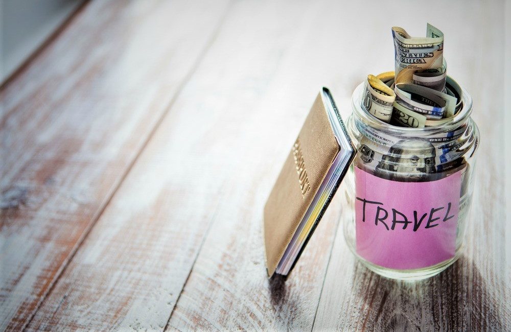  A jar labeled 'travel' filled with money sits beside a passport on a wooden table.